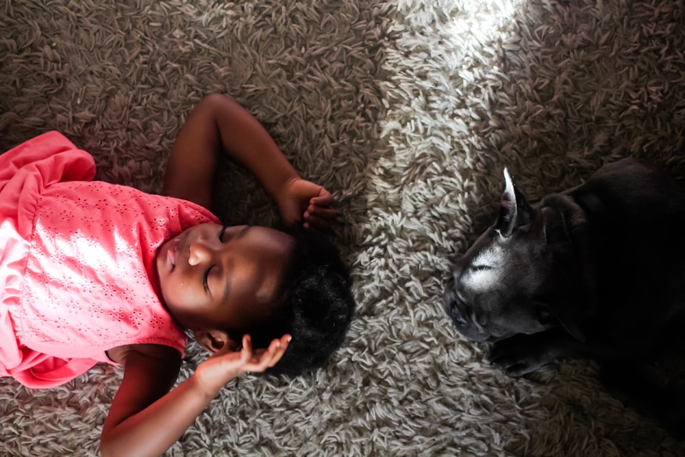 girl laying on rug beside black-short coated animal