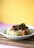 A white bowl filled with spaghetti topped with meatballs in tomato sauce sits on a wooden cutting board. The spaghetti is garnished with chopped herbs for freshness. The background features a warm yellow gradient, adding a vibrant contrast to the image.