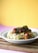 A white bowl filled with spaghetti topped with meatballs in tomato sauce sits on a wooden cutting board. The spaghetti is garnished with chopped herbs for freshness. The background features a warm yellow gradient, adding a vibrant contrast to the image.