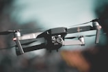A sleek black drone captured in mid-air, with detailed focus on its camera and hardware. The background is a soft blur, emphasizing a smooth bokeh effect and providing a sense of motion.
