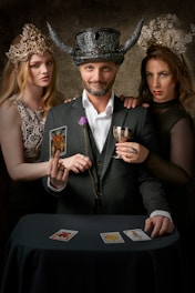 In a dimly lit setting, a man wearing a black ornate horned hat stands between two women adorned with intricate crowns. He holds a goblet and a tarot card, while a single purple rose is tucked into his suit. The women, one on each side, have an enigmatic air, and all three gaze directly ahead. Three tarot cards are spread on a table covered with a dark cloth.