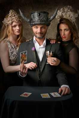 In a dimly lit setting, a man wearing a black ornate horned hat stands between two women adorned with intricate crowns. He holds a goblet and a tarot card, while a single purple rose is tucked into his suit. The women, one on each side, have an enigmatic air, and all three gaze directly ahead. Three tarot cards are spread on a table covered with a dark cloth.