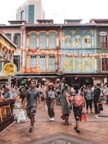 A bustling street scene featuring several people walking and engaging with one another. The area is vibrant, with colorful, traditional-style buildings adorned with lanterns and floral decorations. A variety of shops, including a money changer, are visible along the street.