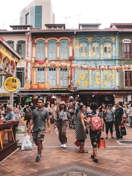 A bustling street scene featuring several people walking and engaging with one another. The area is vibrant, with colorful, traditional-style buildings adorned with lanterns and floral decorations. A variety of shops, including a money changer, are visible along the street.