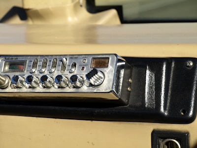 A vintage RadioShack TRC-447 CB radio installed in a vehicle dashboard. The device features various knobs and buttons for controls like volume, squelch, and gain. The dashboard is beige and black with a textured surface and mounting screws.