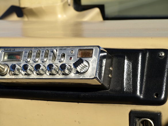 A vintage RadioShack TRC-447 CB radio installed in a vehicle dashboard. The device features various knobs and buttons for controls like volume, squelch, and gain. The dashboard is beige and black with a textured surface and mounting screws.