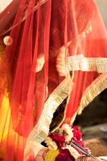 A fabric veil in vibrant red with gold trim partially covers an object or person beneath it. The veil has an intricate design with shimmering gold detailing. Beneath the veil, floral decorations in red and white suggest a festive or ceremonial occasion. A henna design is visible on a hand holding a silver cylindrical object.