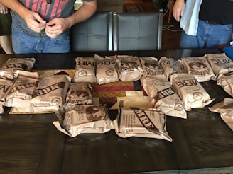 Several packaged MREs are neatly arranged on a dark wooden table. Two individuals, partially visible, stand nearby, one wearing a red shirt and the other in a blue. The table also has a patchwork quilt underneath the MREs.