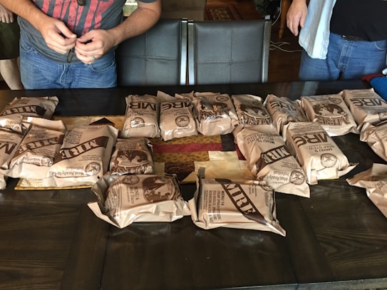 Several packaged MREs are neatly arranged on a dark wooden table. Two individuals, partially visible, stand nearby, one wearing a red shirt and the other in a blue. The table also has a patchwork quilt underneath the MREs.