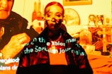 A person is standing in front of a projection that casts various images and text onto them. The projected images include religious symbols, such as a cross, and other household items. Text is visible across the person's face and clothing, creating a mixture of digital and physical elements.