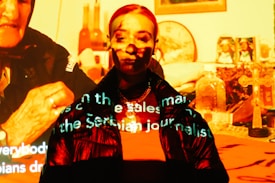 A person is standing in front of a projection that casts various images and text onto them. The projected images include religious symbols, such as a cross, and other household items. Text is visible across the person's face and clothing, creating a mixture of digital and physical elements.