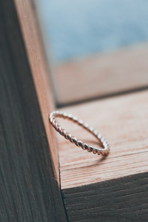 A twisted metal ring is placed on a wooden surface, showcasing its intricate design and craftsmanship. The wood has a natural texture with visible grain, adding warmth to the composition. The background is softly blurred, highlighting the ring as the focal point.