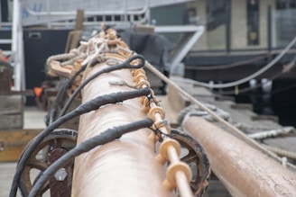 A close-up view of a ship's rigging, showing wooden and metallic elements with ropes intertwined. The background includes blurred structures, possibly parts of a boat or dock. The focus is on the craftsmanship and materials used in maritime equipment.