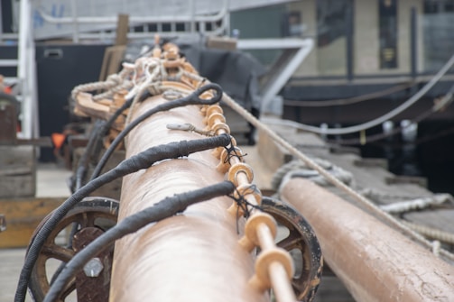 A close-up view of a ship's rigging, showing wooden and metallic elements with ropes intertwined. The background includes blurred structures, possibly parts of a boat or dock. The focus is on the craftsmanship and materials used in maritime equipment.
