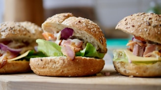 Three freshly made sandwiches with sesame seed whole-grain buns filled with lettuce, bacon, red onion slices, and cheese, placed on a wooden board.