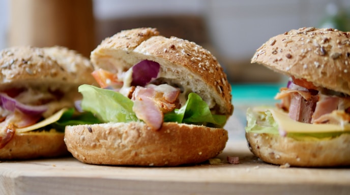 Three freshly made sandwiches with sesame seed whole-grain buns filled with lettuce, bacon, red onion slices, and cheese, placed on a wooden board.