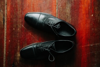 A pair of black leather shoes is placed on a polished wooden floor. The shoes are neatly positioned, and the texture of the wood creates a warm contrast with the smoothness of the leather.