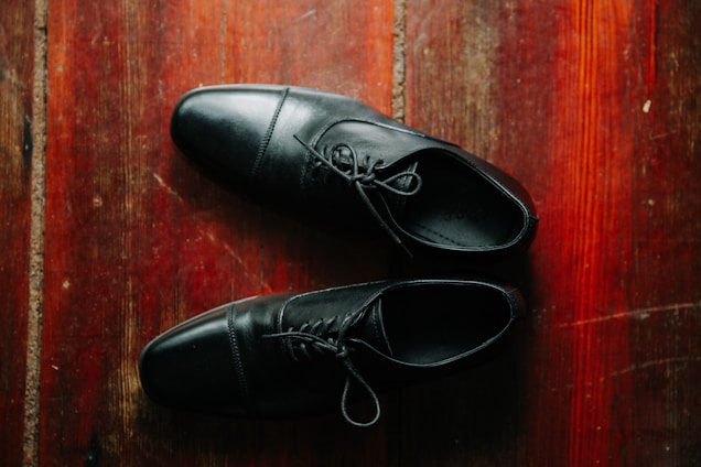 A pair of black leather shoes is placed on a polished wooden floor. The shoes are neatly positioned, and the texture of the wood creates a warm contrast with the smoothness of the leather.