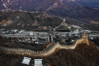 An expansive view of a section of the Great Wall of China winding through rugged terrain with hills and mountains in the background. Below, there is a complex area with parking lots, buildings, and pathways. The landscape is mostly barren, suggesting a dry or winter season.