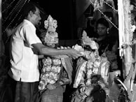 A black and white image depicts a traditional ceremony with a man performing a ritual on a seated married couple. The couple is adorned with ornate clothing and floral garlands, with intricate headdresses. Two women stand behind, observing the ceremony under decorated surroundings.