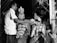 A black and white image depicts a traditional ceremony with a man performing a ritual on a seated married couple. The couple is adorned with ornate clothing and floral garlands, with intricate headdresses. Two women stand behind, observing the ceremony under decorated surroundings.