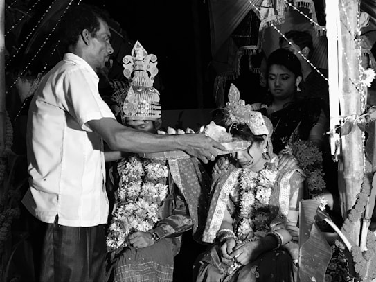 A black and white image depicts a traditional ceremony with a man performing a ritual on a seated married couple. The couple is adorned with ornate clothing and floral garlands, with intricate headdresses. Two women stand behind, observing the ceremony under decorated surroundings.