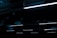 A series of linear fluorescent lights hanging from a ceiling in a dark, industrial setting. The lights cast a minimal blue glow, creating a stark contrast against the otherwise dim environment.