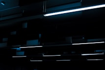 A series of linear fluorescent lights hanging from a ceiling in a dark, industrial setting. The lights cast a minimal blue glow, creating a stark contrast against the otherwise dim environment.