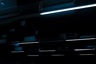 A series of linear fluorescent lights hanging from a ceiling in a dark, industrial setting. The lights cast a minimal blue glow, creating a stark contrast against the otherwise dim environment.