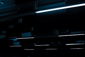A series of linear fluorescent lights hanging from a ceiling in a dark, industrial setting. The lights cast a minimal blue glow, creating a stark contrast against the otherwise dim environment.