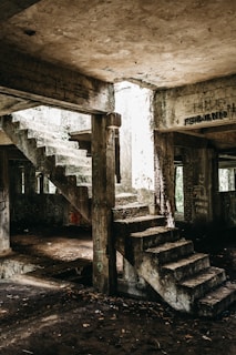 An abandoned concrete structure with two sets of deteriorating stairs and walls covered in graffiti. Natural light pours in from an opening above, illuminating the aged and moss-covered surfaces. The atmosphere is gritty, showcasing signs of neglect and urban decay.