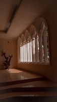 An elegant interior space featuring large, arched windows that allow natural light to stream in, casting defined shadows on the tiled floor. A decorative wooden sculpture is positioned near the windows, adding an artistic element to the scene. The architecture and design suggest a serene and contemplative atmosphere.