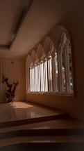 An elegant interior space featuring large, arched windows that allow natural light to stream in, casting defined shadows on the tiled floor. A decorative wooden sculpture is positioned near the windows, adding an artistic element to the scene. The architecture and design suggest a serene and contemplative atmosphere.