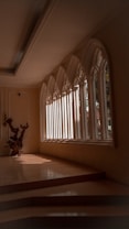 An elegant interior space featuring large, arched windows that allow natural light to stream in, casting defined shadows on the tiled floor. A decorative wooden sculpture is positioned near the windows, adding an artistic element to the scene. The architecture and design suggest a serene and contemplative atmosphere.