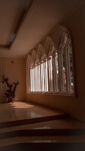 An elegant interior space featuring large, arched windows that allow natural light to stream in, casting defined shadows on the tiled floor. A decorative wooden sculpture is positioned near the windows, adding an artistic element to the scene. The architecture and design suggest a serene and contemplative atmosphere.