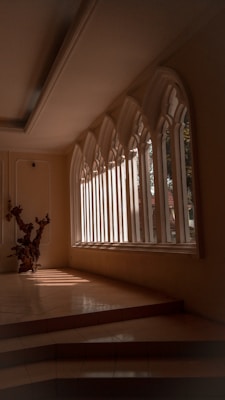 An elegant interior space featuring large, arched windows that allow natural light to stream in, casting defined shadows on the tiled floor. A decorative wooden sculpture is positioned near the windows, adding an artistic element to the scene. The architecture and design suggest a serene and contemplative atmosphere.