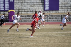 A group of female athletes are playing lacrosse on a grassy field. One player wearing a red uniform is in the foreground holding a lacrosse stick, appearing to be in motion. Several players in white uniforms are in the background, chasing the player in red. A referee and a coach are also visible. The background features a stone wall with purple banners listing years of achievement.