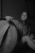 A person with long hair sits in a relaxed pose, gently touching a large drum. The setting is dimly lit, creating a calm and introspective atmosphere. The individual is wearing a striped outfit, and the composition is captured in black and white.