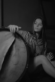 A person with long hair sits in a relaxed pose, gently touching a large drum. The setting is dimly lit, creating a calm and introspective atmosphere. The individual is wearing a striped outfit, and the composition is captured in black and white.