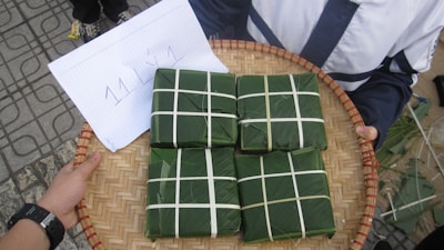 A hand-woven bamboo tray holds four square-shaped packages wrapped in green leaves, resembling traditional food items. Each package is tied with white string in a grid pattern. A paper with handwritten text lies next to the packages.