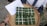 A hand-woven bamboo tray holds four square-shaped packages wrapped in green leaves, resembling traditional food items. Each package is tied with white string in a grid pattern. A paper with handwritten text lies next to the packages.