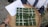 A hand-woven bamboo tray holds four square-shaped packages wrapped in green leaves, resembling traditional food items. Each package is tied with white string in a grid pattern. A paper with handwritten text lies next to the packages.