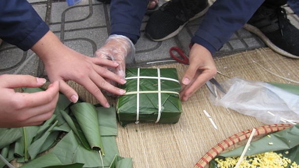 Several hands are involved in wrapping a square package using large green leaves and white string. The activity takes place on a woven mat, and other leaves and objects like scissors and a woven basket with yellow contents are visible.