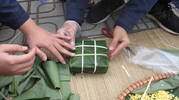 Several hands are involved in wrapping a square package using large green leaves and white string. The activity takes place on a woven mat, and other leaves and objects like scissors and a woven basket with yellow contents are visible.