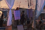 In a bohemian outdoor setting, dreamcatchers and colorful fabric hang from lines with a warm, dusky sky in the background. Various people stand and sit amidst this artistic arrangement, surrounded by white drapes and decorative textiles.