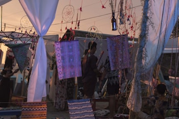 In a bohemian outdoor setting, dreamcatchers and colorful fabric hang from lines with a warm, dusky sky in the background. Various people stand and sit amidst this artistic arrangement, surrounded by white drapes and decorative textiles.