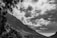 A dramatic black and white landscape photograph depicting a mountainous terrain. Large trees frame the scene on the left, and rugged peaks rise towards a sky filled with textured clouds.