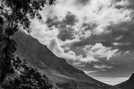 A dramatic black and white landscape photograph depicting a mountainous terrain. Large trees frame the scene on the left, and rugged peaks rise towards a sky filled with textured clouds.