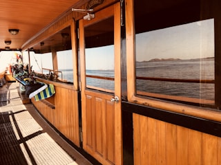 Wooden paneling covers the side of a boat deck with a door leading inside. A row of scuba gear and other equipment is lined up on a bench, indicating a preparation area for diving. The sea and distant islands are visible through the glass and from the deck, suggesting a calm marine environment.