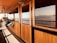 Wooden paneling covers the side of a boat deck with a door leading inside. A row of scuba gear and other equipment is lined up on a bench, indicating a preparation area for diving. The sea and distant islands are visible through the glass and from the deck, suggesting a calm marine environment.